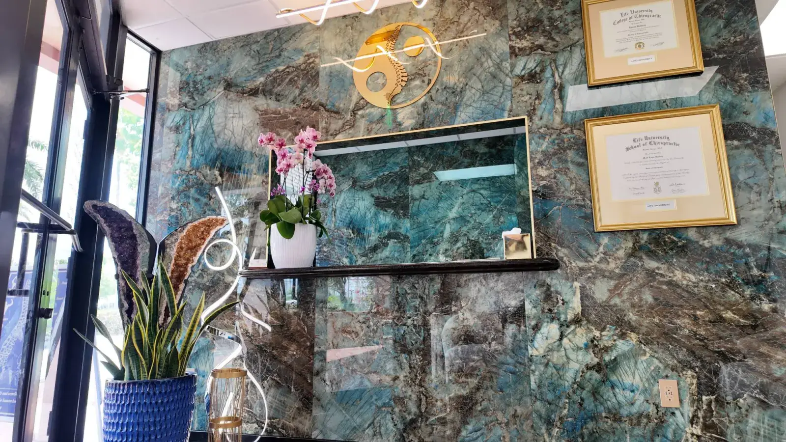 The front desk at Luxury Chiropractic in North Miami Beach, with the practice's yin yang spine mark and the doctors' Life University diplomas on the marble wall.