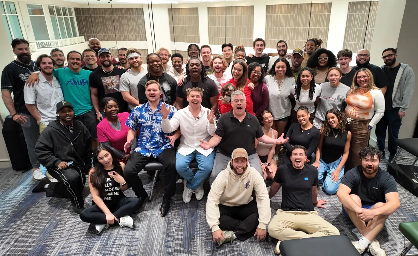 Podcast A large seminar class of practitioners gathered for a photograph, several seated on the floor, portable tables stacked at the edges of the room.