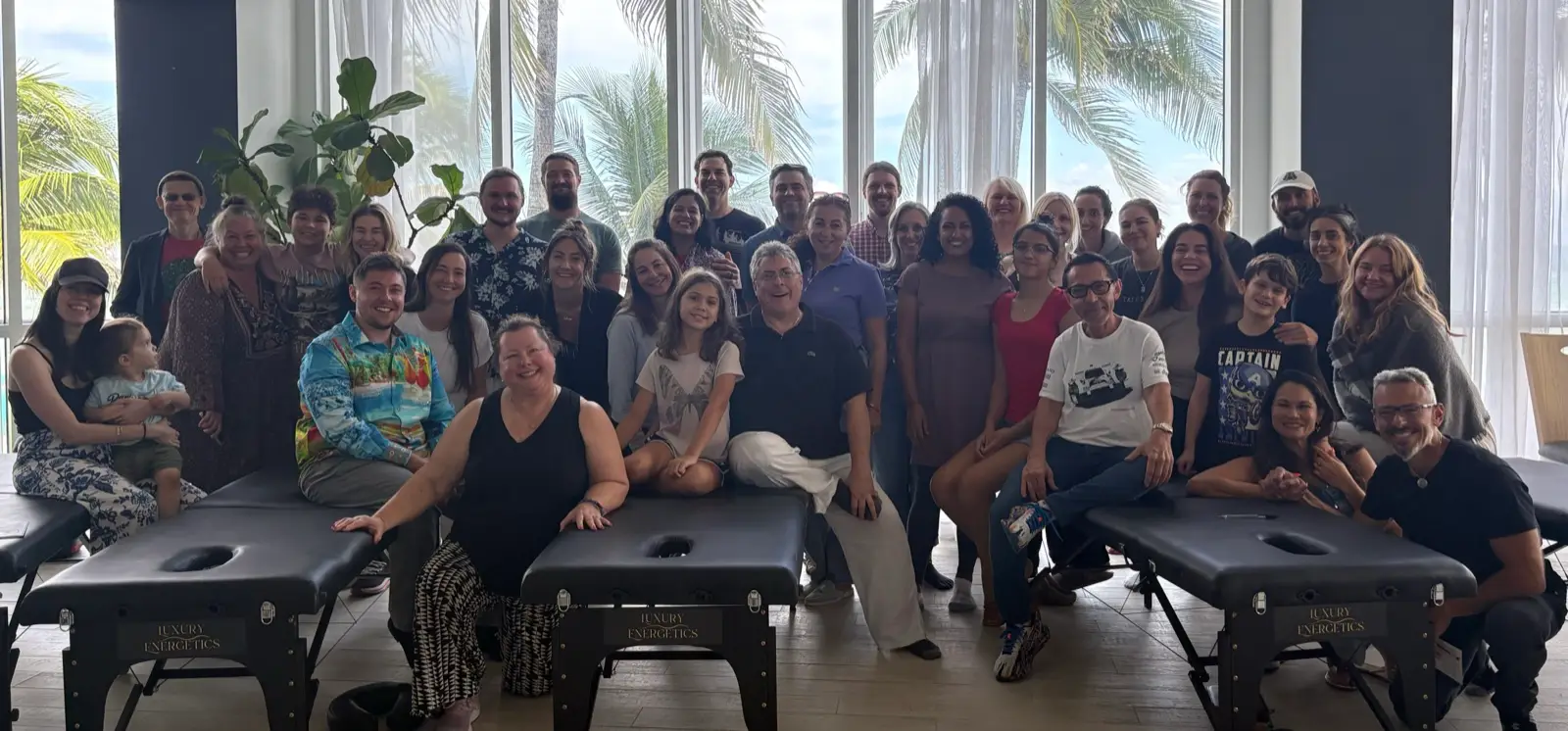Podcast Practitioners and families gathered beside Luxury Energetics tables in a seminar room with floor to ceiling windows and palm trees outside.