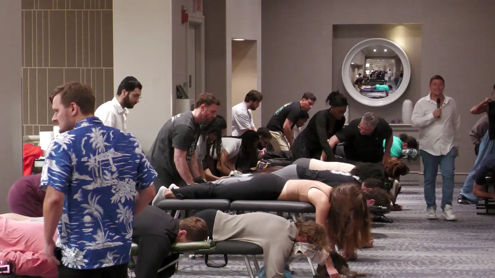 Podcast Rows of portable tables on a seminar floor, practitioners working in pairs with their hands on patients spines while an instructor speaks into a microphone.